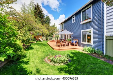 Wooden Walkout Deck In The Backyard Garden Of Blue Siding House. Furnished With Patio Table Set With Umbrella