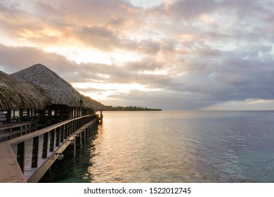 A wooden walkway with thatched roof bungalows leads out into the lagoon on the island of Moorea in French Polynesia at sunset; ocpy space - Powered by Shutterstock