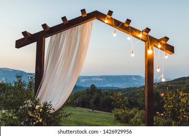 Wooden wedding arch with white cloth and light bulbs outdoors with amazing mountain view on background. Night ceremony in twilight.