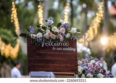 Wooden welcome board sign with a beautiful flower decoration, standing in front of wedding entrance.