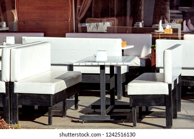 A wooden white table with white sofas on black legs against the background of the facade with windows of an outdoor cafe with empty seats on a sunny day.