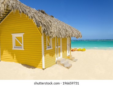 Wooden Yellow Hut On The Beach Covered With Thatch Against Colorful Kayaks, Blue Sky And Azure Water, Caribbean Islands 