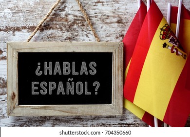 a wooden-framed chalkboard with the question hablas espanol? do you speak Spanish? written in Spanish, and some flags of Spain against a rustic wooden background