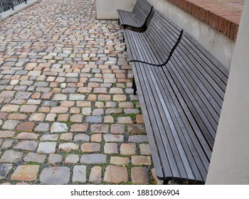 Wood-paneled Benches By A Bright Plaster Wall In A Park Sunk With Leaves And Creepers Other Clean And Tidy Around. Cubes Paving Granite Gray. Brick Attic Wall. Multiple Benches In A Row Behind Gravel,