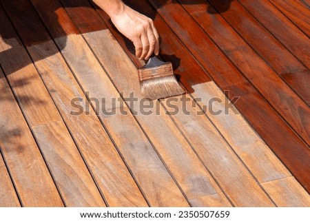 Woodworker refreshing wooden floor, worker's hand is applying decking oil on deck with a painting brush after sanding