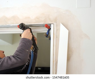 Woodworker Using A Compressed Air Nailer Tool To Insert Nails Into The Flashing Frame Of A New White Door