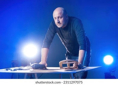 Woodworker at work. Man near table with boards. Owner home carpentry workshop. Artisan guy looks into camera. Woodworker with power tool for woodworking. Adult woodworker man on blue background - Shutterstock ID 2311297075