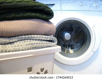 Woolen Items Are Lying On A White Laundry Basket And Waiting For Washing Against The Background Of The Washing Machine