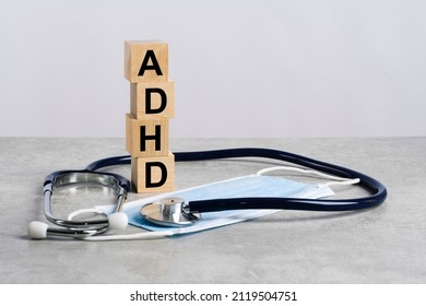 Word Adhd Is Made Of Wooden Cubes On A Gray Background With Stethoscope And Medicine Mask. Medical Concept Of Prevention And Treatment. Adhd - Short For Attention Deficit Hyperactivity Syndrome.