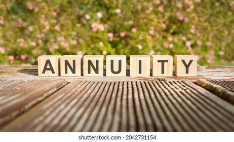 The Word Annuity Is Written On Wooden Cubes. The Blocks Are Placed On An Old Wooden Board Illuminated By The Sun. In The Background Is A Brightly Blooming Shrub