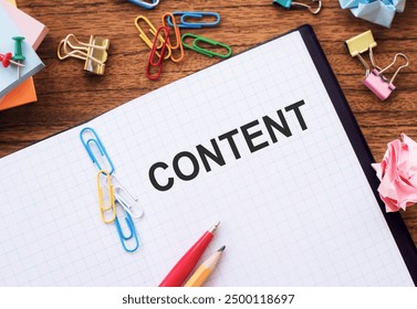 The word CONTENT written on a white grid notebook on a wooden table. There are various office supplies such as paper clips, sticky notes, pencils, and crumpled paper nearby. - Powered by Shutterstock