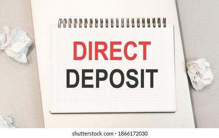 The word direct deposit is written in a notebook that sits on a gray desktop along with a laptop.