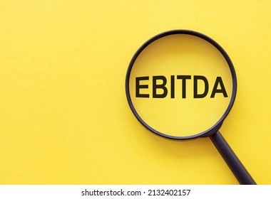 The word EBITDA - Earnings Before Interest, Taxes, Depreciation and Amortization, is written on a magnifying glass on a yellow background.