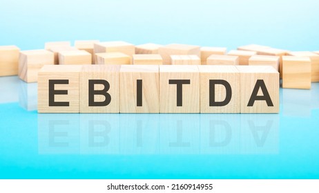 word ebitda made with wood building blocks. text is written in black letters and is reflected in the mirror surface of the table, blue background, business concept
