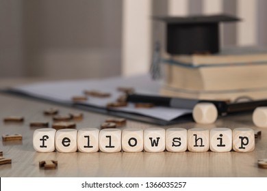 Word FELLOWSHIP composed of wooden dices. Black graduate hat and books in the background. Closeup