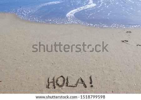 The word hello in Spanish with an exclamation point (Hola!) on a golden sandy beach with blue ocean waves rolling in from above.  Peaceful, tranquil, tropical, vacation concepts.