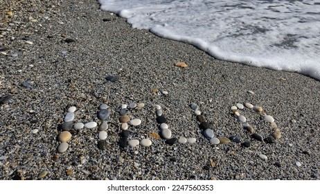 The word Hello is written with small stones on the beach in Nerja. The word is written in capital letters. Waves are crashing in on the beach. The sand is grey and wet. - Powered by Shutterstock