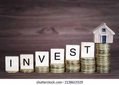 Word INVEST made from wooden letter blocks and a miniature house placed on ascending coins stacks. Creative home investment concept.