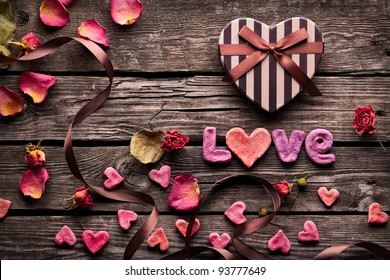Word Love with Heart shaped Valentines Day gift box on old vintage wooden plates. Sweet holiday background with rose petals, small hearts, curved ribbon.