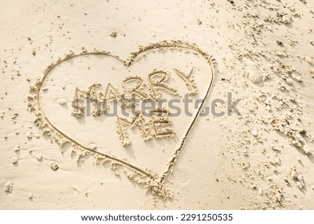 The word MARRY ME is writting on the sand inside heart shaped sand writing. drawing on the sand. writing with sands on the beach.
