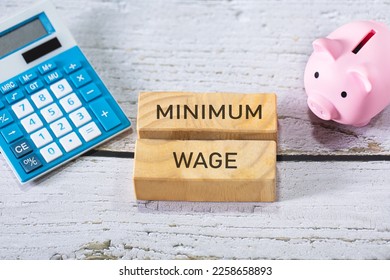The word minimum wage inscribed in wooden blocks. A piggy bank and a calculator on a wooden table in the composition. Economy.
