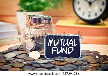 Word Mutual Fund on mini chalkboard and coin in the jar with blurred background of books, green plant and clock. Financial Concept.