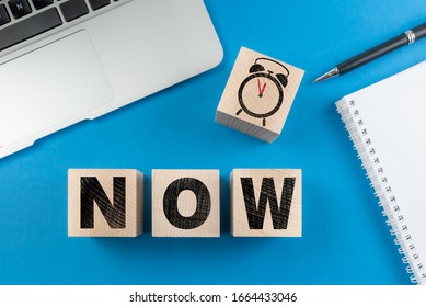 word NOW and alarm clock on wooden cubes on blue desk background with notepad and laptop computer