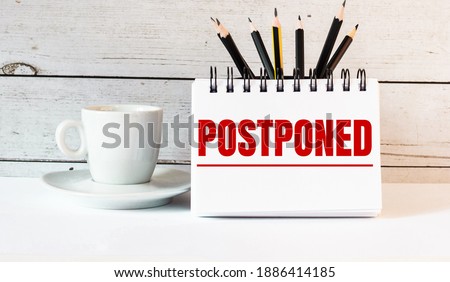 The word POSTPONED is written in a white notepad near a white cup of coffee on a light background