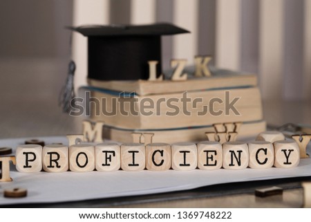 Word PROFICIENCY composed of wooden dices. Black graduate hat and books in the background. Closeup
