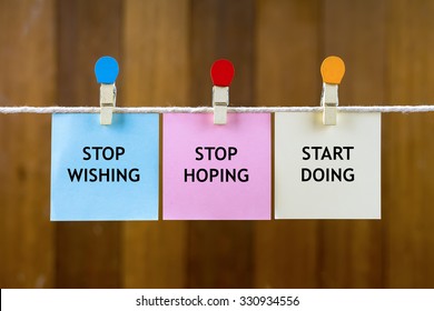 Word quotes of STOP WISHING, STOP HOPING, START DOING on colorful sticky papers hanging by a rope against blurred wooden background.