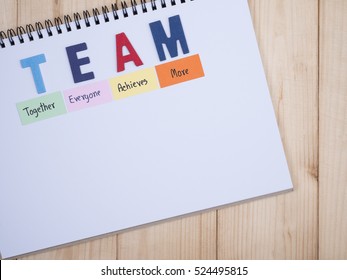 Word spelling TEAM and handwriting together, everyone, achieve, more on notebook with wood background (Business Concept)