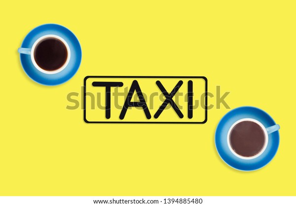 Word taxi in frame of black color cut\
out of black paper near full blue cups of hot chocolate and coffee\
on yellow background. Top view. Concept of\
journey