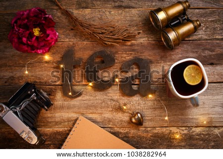 The word "tea" spelled using brown wooden letters on the wooden table. A cup of tea with a lemon. Binoculars, old camera, tea sieve, notebook, red peony flower, garland with a yellow bulbs.
