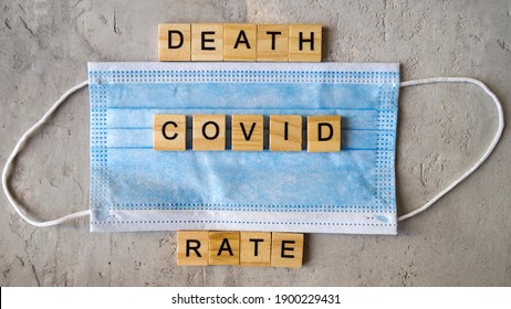 Words DEATH RATE COVID Lined With Wooden Letters On A Background Of Blue Surgical Mask.