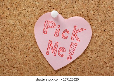 The words Pick Me on a pink heart shaped post it note pinned to a cork notice board