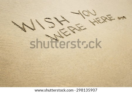 Words wish you were here written in the sand on the beach.