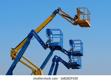 Work Aerial Platforms Against Blue Cloudless Sky. One Cherry Picker Yellow Among Three Blue