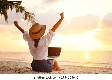 Work from anywhere. Rear view of young woman, female freelancer in straw hat working on laptop, keeping arms raised and cellebrating success while sitting on the tropical sandy beach at sunset