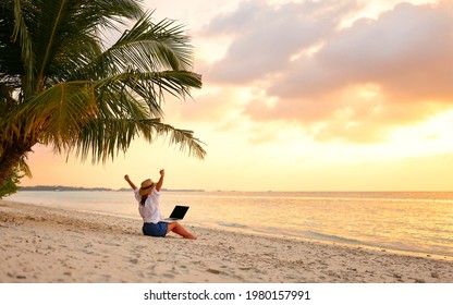 Work From Anywhere. Rear View Of Young Woman, Female Freelancer In Straw Hat Working On Laptop, Keeping Arms Raised And Cellebrating Success While Sitting On The Tropical Sandy Beach At Sunset