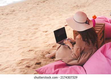 Work And Business Travel Concept. Rear View Of Young Adult Southeast Asian Woman Using Laptop Computer At The Sand Beach Pink Seaside Luxury Cafe. Freelance Lifestyle On Holidays.