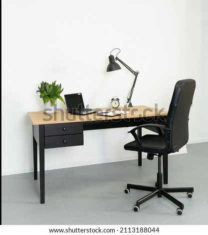 A work desk with a wooden top and a black base. On the desk there is a room flower, a laptop, a lamp, an alarm clock, a notebook and a pen. Next to the desk is a black office chair. Workplace in the o