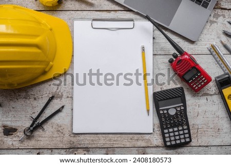 work environment with a laptop, a yellow hard hat, calculator, level, screwdriver, and folding ruler on a well-used wooden surface, suggesting a construction or engineering context.
