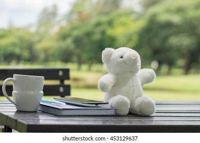 Work In The Garden Concept, Coffee Cup, Book, Smart Phone And White Bear On Wooden Table Under The Spring Sunlight In A Park. Using As Background Or Wallpaper Nature Template