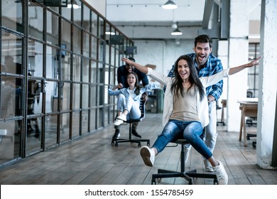 Work Hard Play Hard. Four Young Cheerful Business People In Smart Casual Wear Having Fun While Racing On Office Chairs And Smiling.