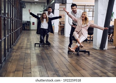 Work Hard Play Hard. Four Young Cheerful Business People In Formal Wear Having Fun While Racing On Office Chairs And Smiling