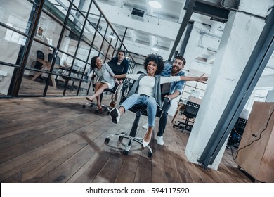 Arbeite fleißig beim Spielen!  Vier junge, fröhliche Geschäftsleute in intelligenter, ungezwungener Kleidung, die Spaß beim Rennen auf Bürostühlen und Lächeln haben