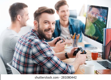 Work Hard Play Hard. Three Young Men Playing Computer Games While Sitting At The Desk In The Office