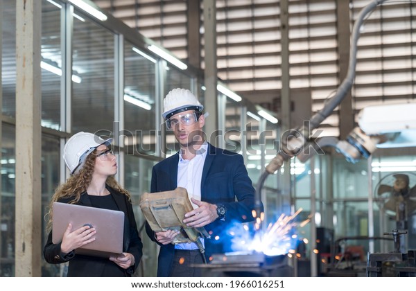 Work in\
a Heavy Industry Manufacturing Factory. Industrial man and woman\
engineer with laptop in control robot of\
factory.