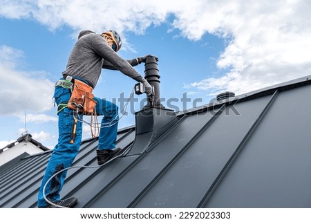 Work at heights. A tinman with tool belt and climbing harness on the roof is working with a cordless drill on the chimney sheeting.