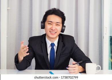 Work from home, Asian young businessman wear suit video conference call or facetime he smiling sitting on desk wearing headphones and raise his hand to explain the job he looking to camera - Powered by Shutterstock